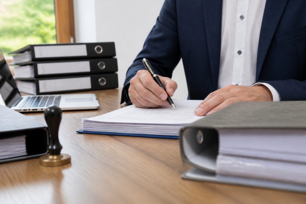 Geschäftsperson unterzeichnet rechtliche Dokumente an einem Schreibtisch mit mehreren Ordnern und Laptop im Hintergrund, natürliche Beleuchtung